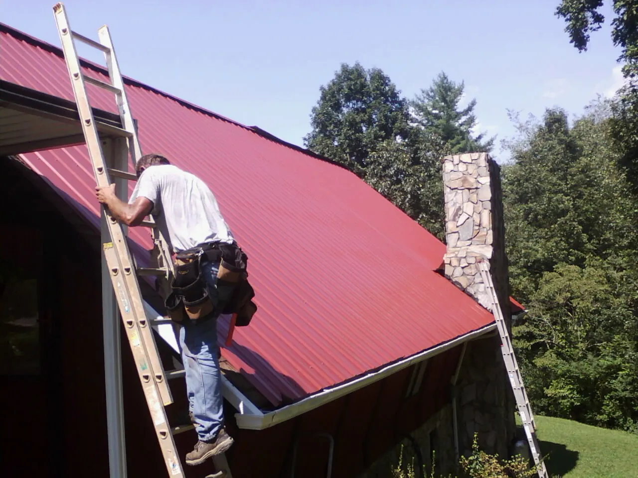 Roofer on a ladder beside a red metal roof and stone chimney — Soffit & Fascia Repair by Roofing Brothers in Palmyra